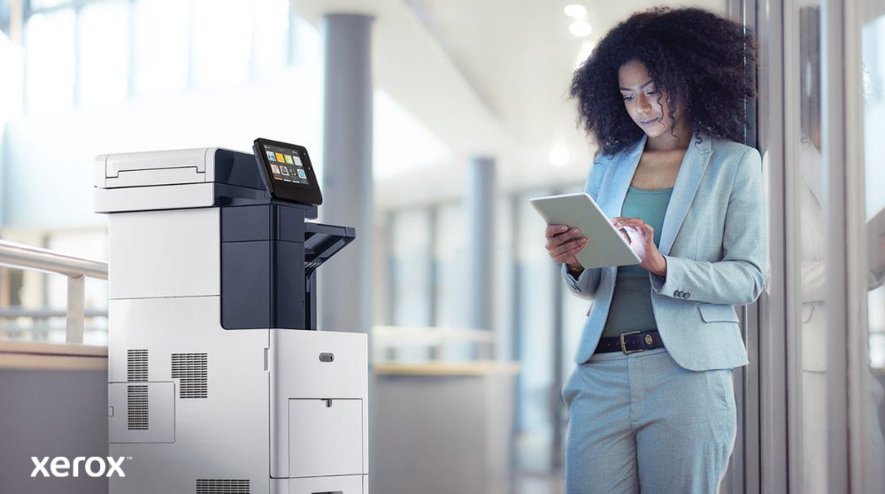 Une personne se tient à côté d'une imprimante multifonction Xerox VersaLink B605, regardant et interagissant avec une tablette, illustrant comment ConnectKey permet une interaction mobile avec l'imprimante.
