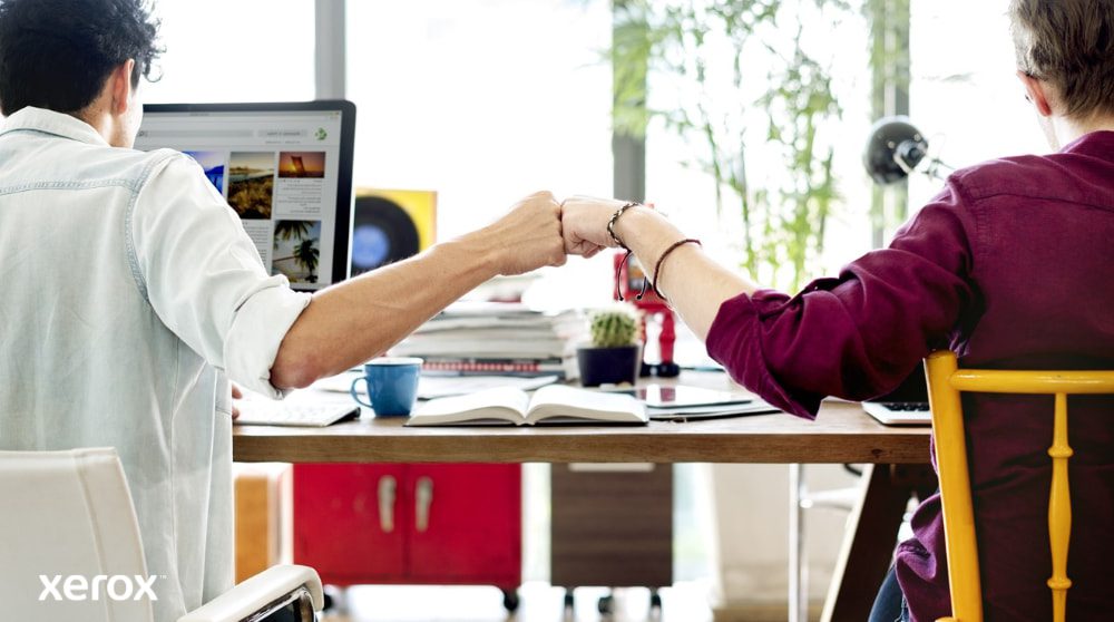 Xerox® Smart Start : travail d'équipe et collaboration Deux personnes assises à un bureau, dos à la caméra, se serrent le poing, symbole du travail d'équipe et d'une collaboration réussie. L'une d'elles regarde un écran d'ordinateur.