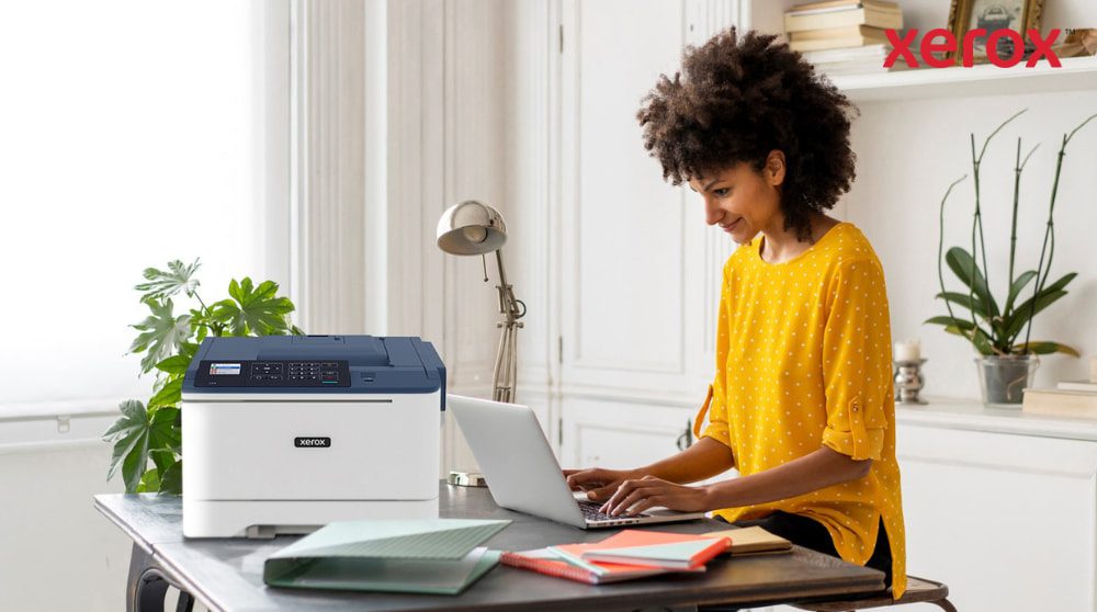 Xerox® DocuShare® Go : productivité pour le travail hybride Une personne aux cheveux bouclés sourit tout en tapant sur un ordinateur portable à un bureau, avec une imprimante compacte Xerox C310 positionnée à côté de l'ordinateur portable, présentant une configuration de bureau à domicile moderne pour la productivité.
