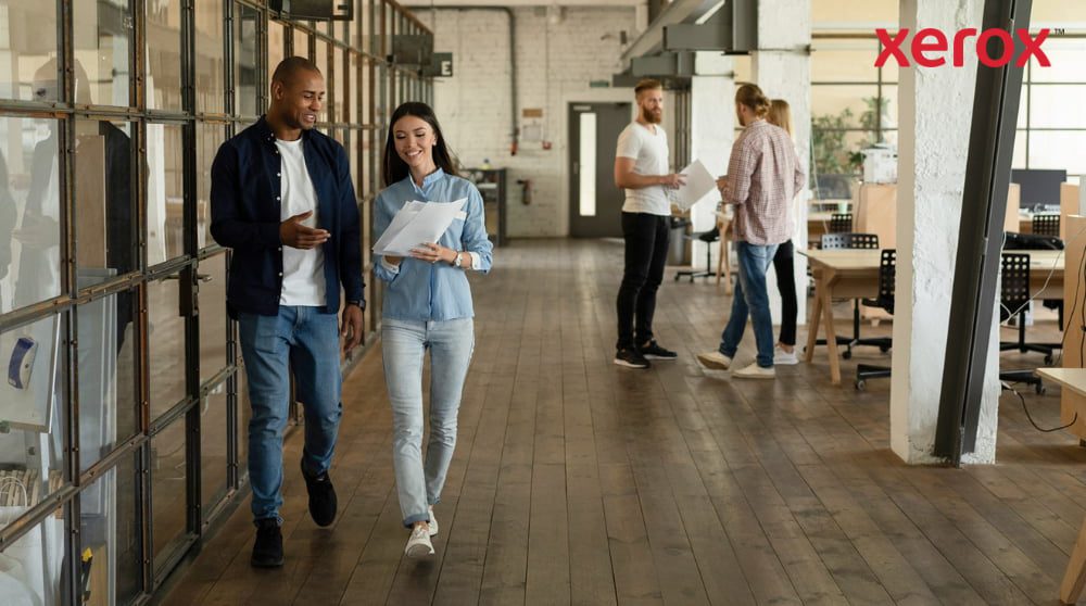 Expérience d'impression et de numérisation Xerox® : dynamiser la collaboration sur le lieu de travail Deux collègues marchent dans un couloir d'un bureau moderne et ouvert, tenant des documents et souriant, avec d'autres collègues visibles en arrière-plan, représentant un lieu de travail dynamique et collaboratif.